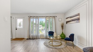 A modern living room with a wooden floor, a round coffee table, two blue chairs, a glass table, and a door leading outside. - Hot Tub Haven w/ King Bed + More (Greater Sudbury)