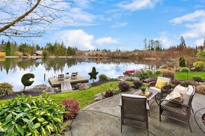 A lakeside patio with outdoor furniture, a small deck, and a reflection of the surrounding trees and buildings.