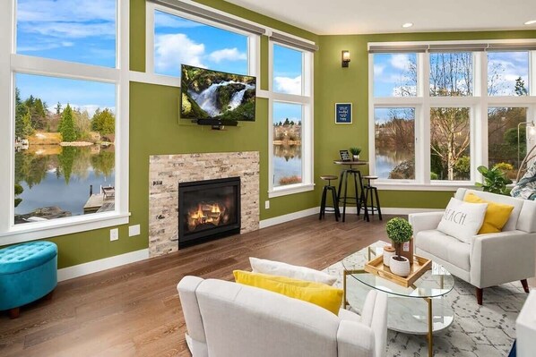 A spacious living room with a fireplace, large windows offering a view of a lake, and a comfortable seating area.