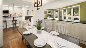 Una cucina moderna con mobili bianchi, un'area pranzo con tavolo in legno apparecchiata per sei persone e vista all'esterno tramite ampie finestre.