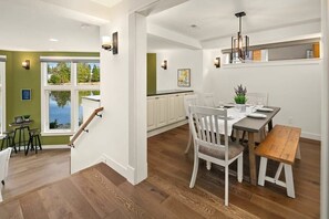 Een moderne keuken met witte kasten, een eethoek met een houten tafel en stoelen, en uitzicht op de omgeving door grote ramen.