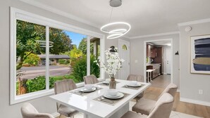 Ein moderner Essbereich mit einem weißen Tisch, gedeckt für sechs Personen, großen Fenstern mit Blick ins Freie und einer durch einen offenen Durchgang einsehbarer Küche.