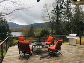 A deck with outdoor furniture and a grill, overlooking a lake and mountains.