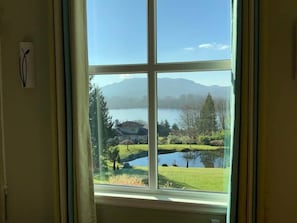 Une fenêtre donnant sur un lac, une maison et des montagnes au loin.