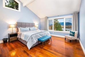 A bedroom with a bed, bedside tables, a bench, a chair, and a large window with a view of trees and mountains.