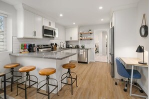 A modern kitchen with white cabinets, a stainless steel refrigerator, a microwave, and a coffee machine. There are three wooden stools and a blue office chair.