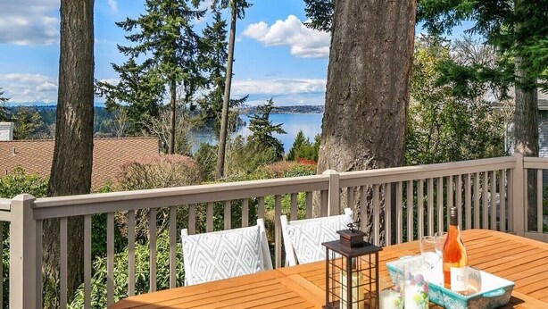 A wooden deck with a table set for two, overlooking a lake with trees and a distant cityscape. - Family Retreat w Lake & Mt & City View, Gym, MI (Mercer Island)