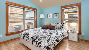 A bedroom with a large bed, two windows, a nightstand, and a lamp.