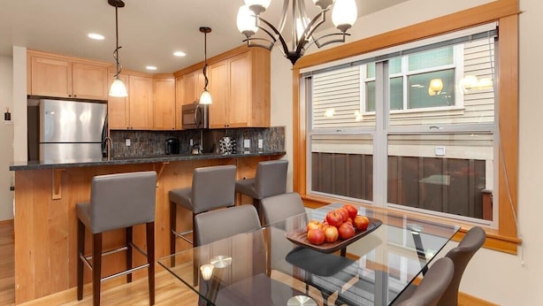 A modern kitchen with wooden cabinets, a stainless steel refrigerator, a glass dining table with chairs, and a chandelier.