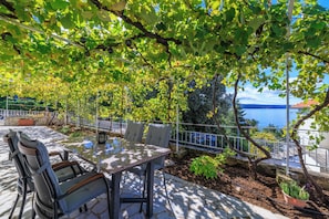 Um pátio externo com mesa e cadeiras, rodeado por vegetação e vista para o mar.