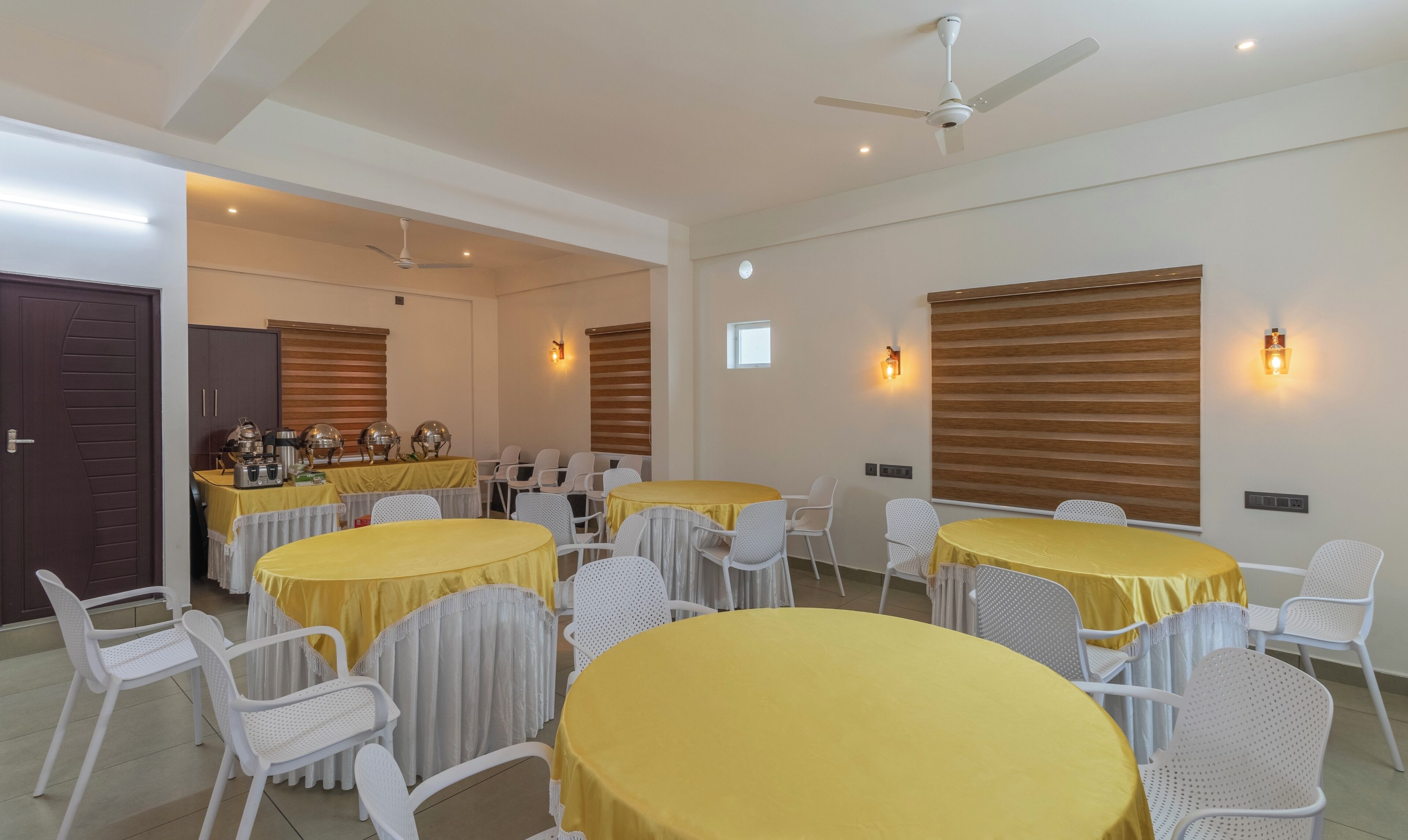 A dining area with round tables covered in yellow tablecloths, white chairs, and ceiling fans.