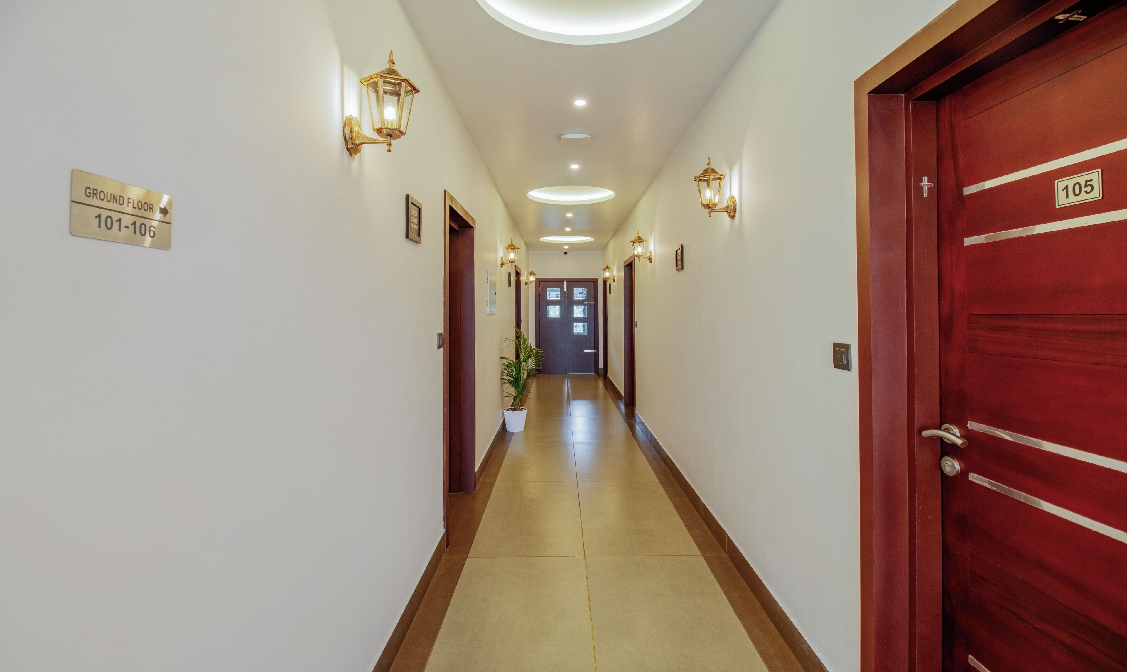 Corredor de hotel com placa indicando quartos do andar térreo 101 a 106, um vaso de planta e uma porta vermelha com o número 105.