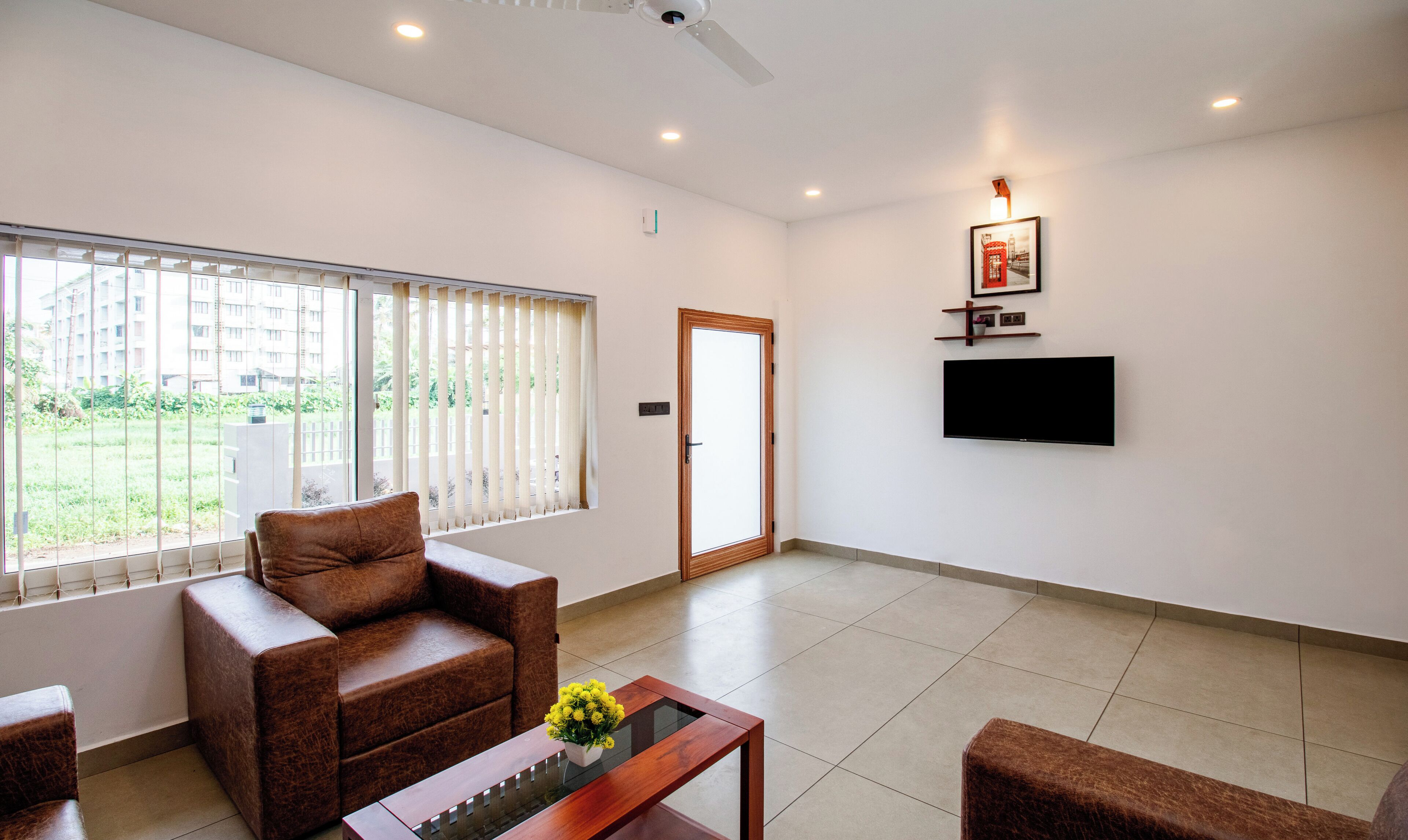A modern living room with a sofa, a coffee table with flowers, a flat-screen TV mounted on the wall, and a large window with blinds.