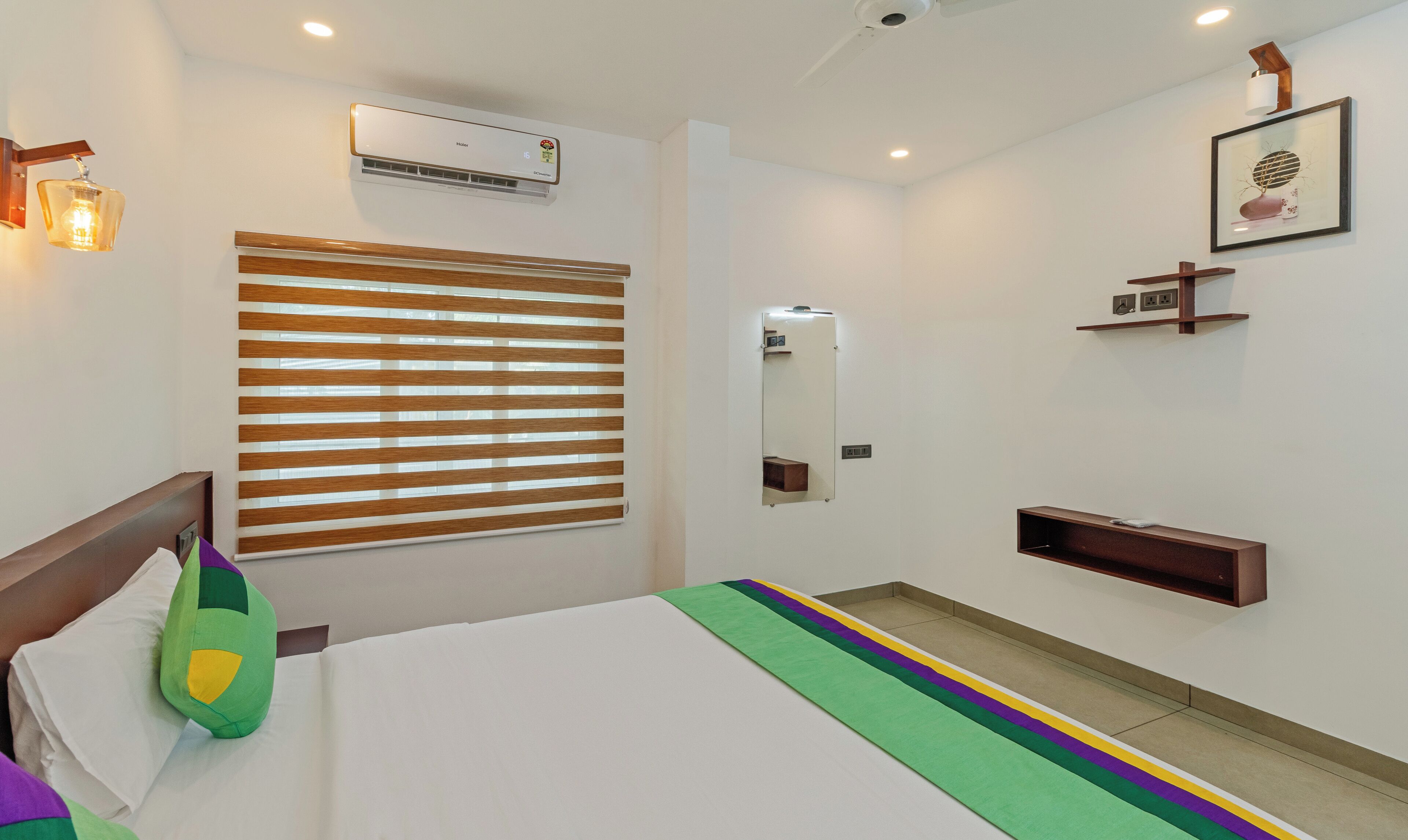 A hotel room with a bed, a window with blinds, an air conditioning unit, a bedside table, a mirror, and a wall-mounted shelf.