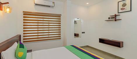 A hotel room with a bed, a window with blinds, an air conditioning unit, a bedside table, a mirror, and a wall-mounted shelf.