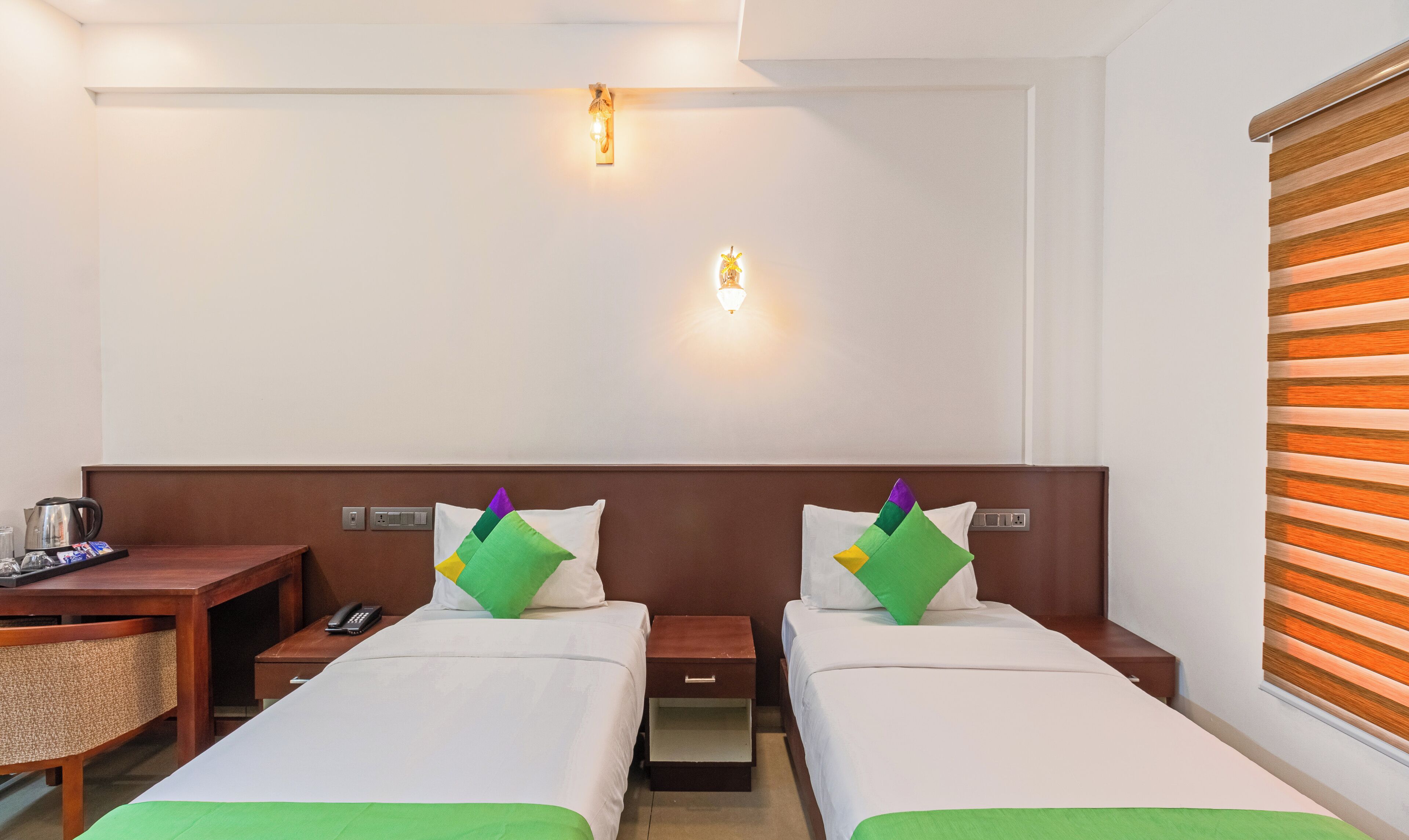 A hotel room with two single beds, a wooden nightstand with a phone, a small table with a kettle and a tray, and a wall-mounted light fixture.
