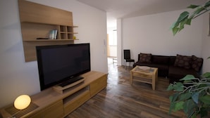 Ein modernes Wohnzimmer mit Flachbildfernseher, Holzmöbeln, einer Couch mit gemusterten Kissen und einer Pflanze.