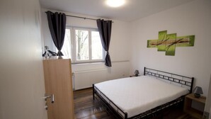 Ein Schlafzimmer mit Holzboden, einem Bett mit weißer Matratze, einem Nachttisch mit Lampe, einem Fenster mit Vorhängen und einem Heizkörper.