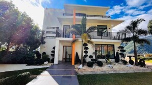 Una casa moderna de dos pisos con balcón, rodeada de jardines y árboles bien cuidados.