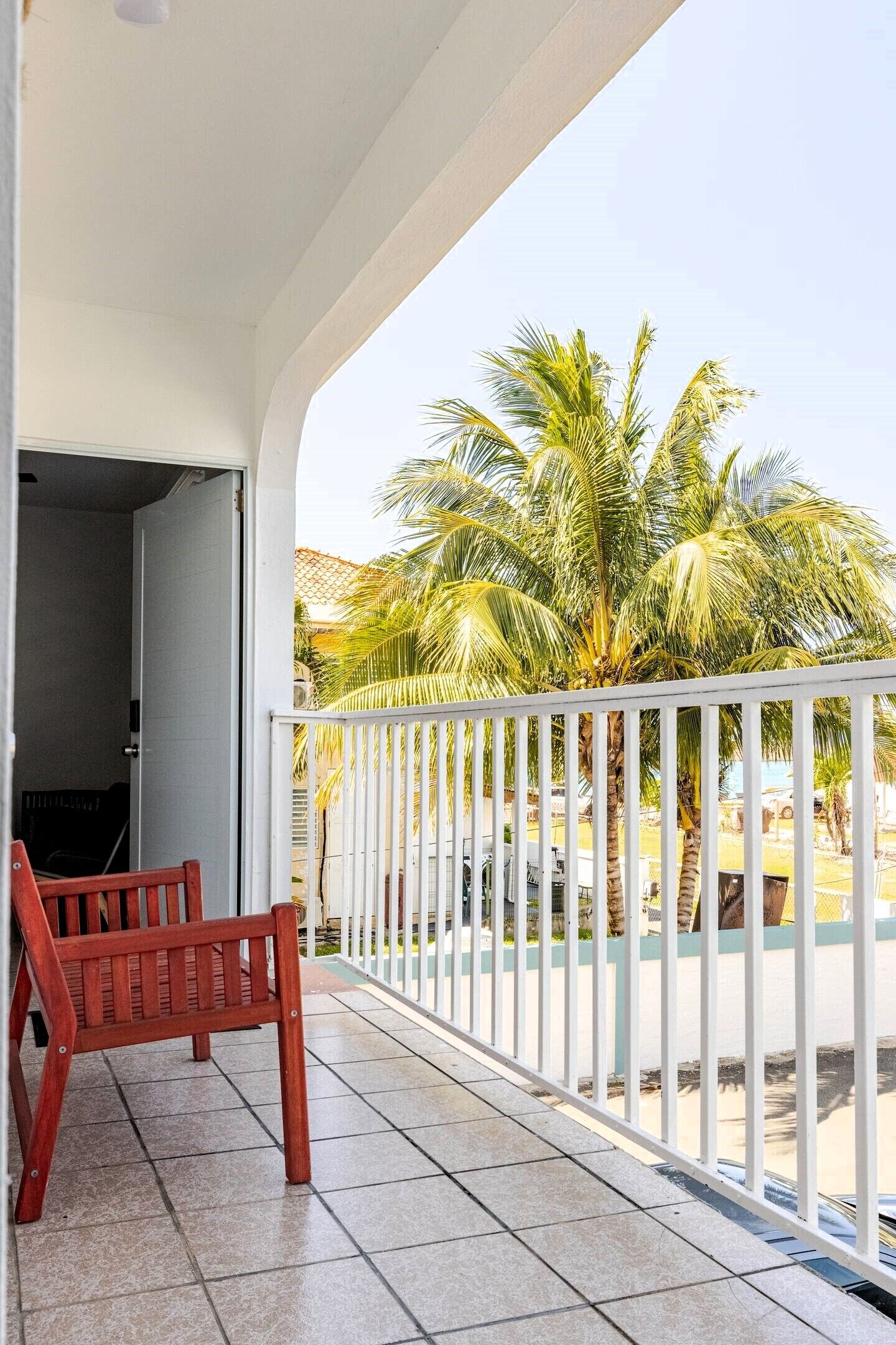 A balcony with a white railing, a wooden bench, and a view of palm trees.