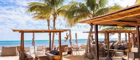 A beachside lounge with wooden furniture, palm trees, and a view of the ocean.
