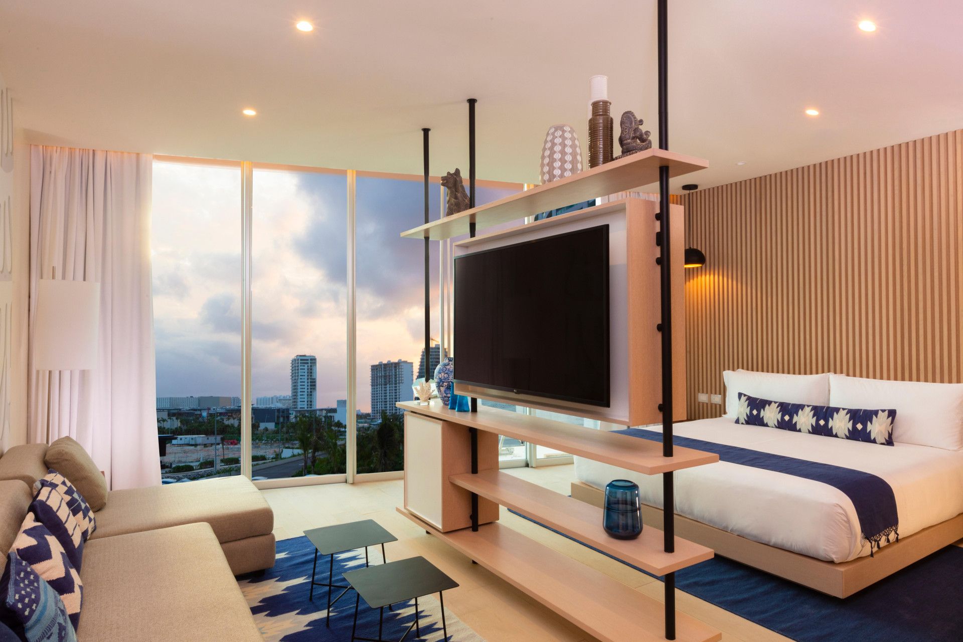A modern hotel room with a large TV, a sofa, a bed, and a city view.