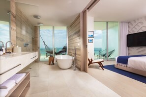 Une chambre d’hôtel moderne dotée d’une grande baignoire, d’une douche séparée, d’un lit avec télévision et d’une vue sur l’océan.