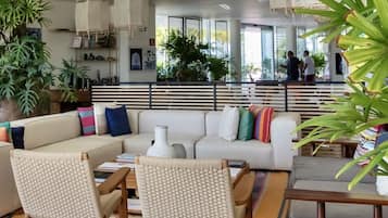 A modern living room with a white sofa, wicker chairs, a coffee table, and a striped rug. There are plants, a bookshelf, and a staircase in the background.