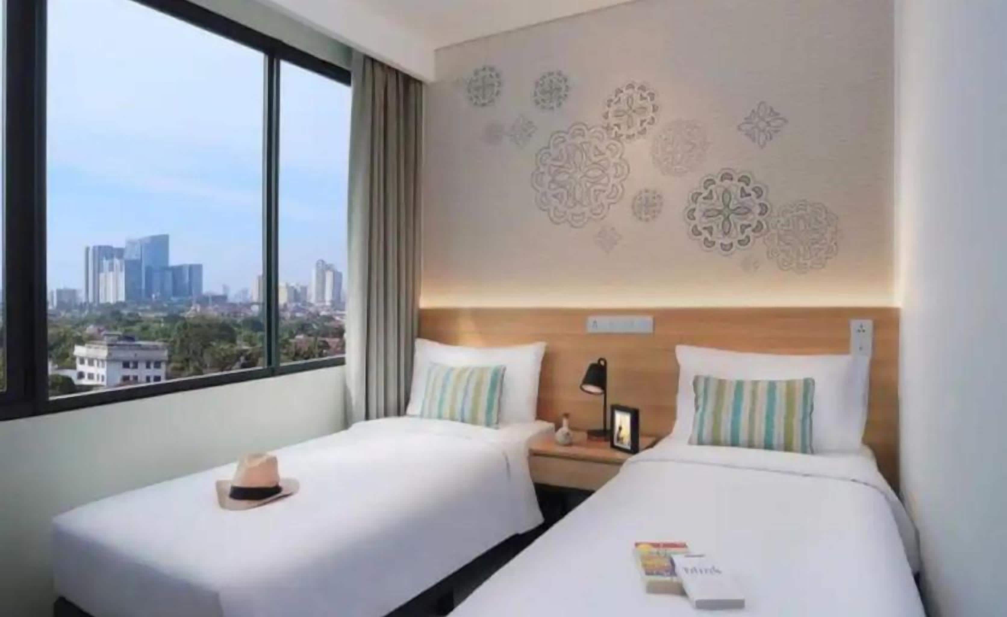 Ein Hotelzimmer mit zwei Betten, einem Nachttisch, einem Fenster mit Stadtblick und dekorativen Wandmustern.