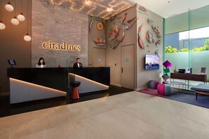 A reception desk with a 'Citadines' sign, a modern interior with abstract wall art, and a seating area with colorful stools. - Citadines Gatot Subroto Jakarta (JAKARTA)