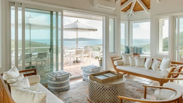 A spacious room with a large sliding glass door leading to an outdoor area with a table and chairs. There is a wooden bench with cushions and a small round table with a tray. - Villa Magli (Christiansted)