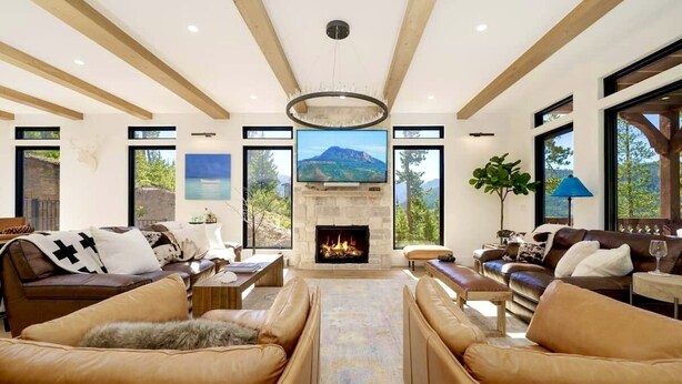 A spacious living room with a fireplace, leather sofas, and large windows offering a mountain view. - Swiss Chalet: Hot Tub, EV Charger, Lake Views (Grand Lake)