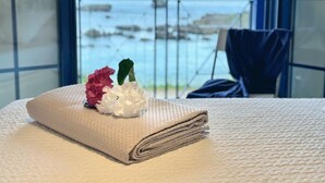 Una cama bien hecha con una cubierta blanca texturizada, una flor rosa y una flor blanca en un asiento junto a la ventana con vista a un cuerpo de agua.