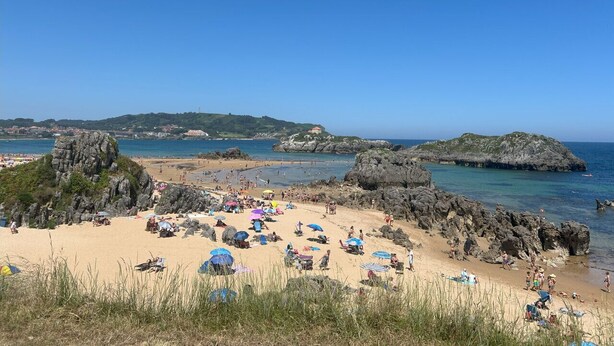 Una playa con formaciones rocosas, cielo azul despejado y gente disfrutando del sol.