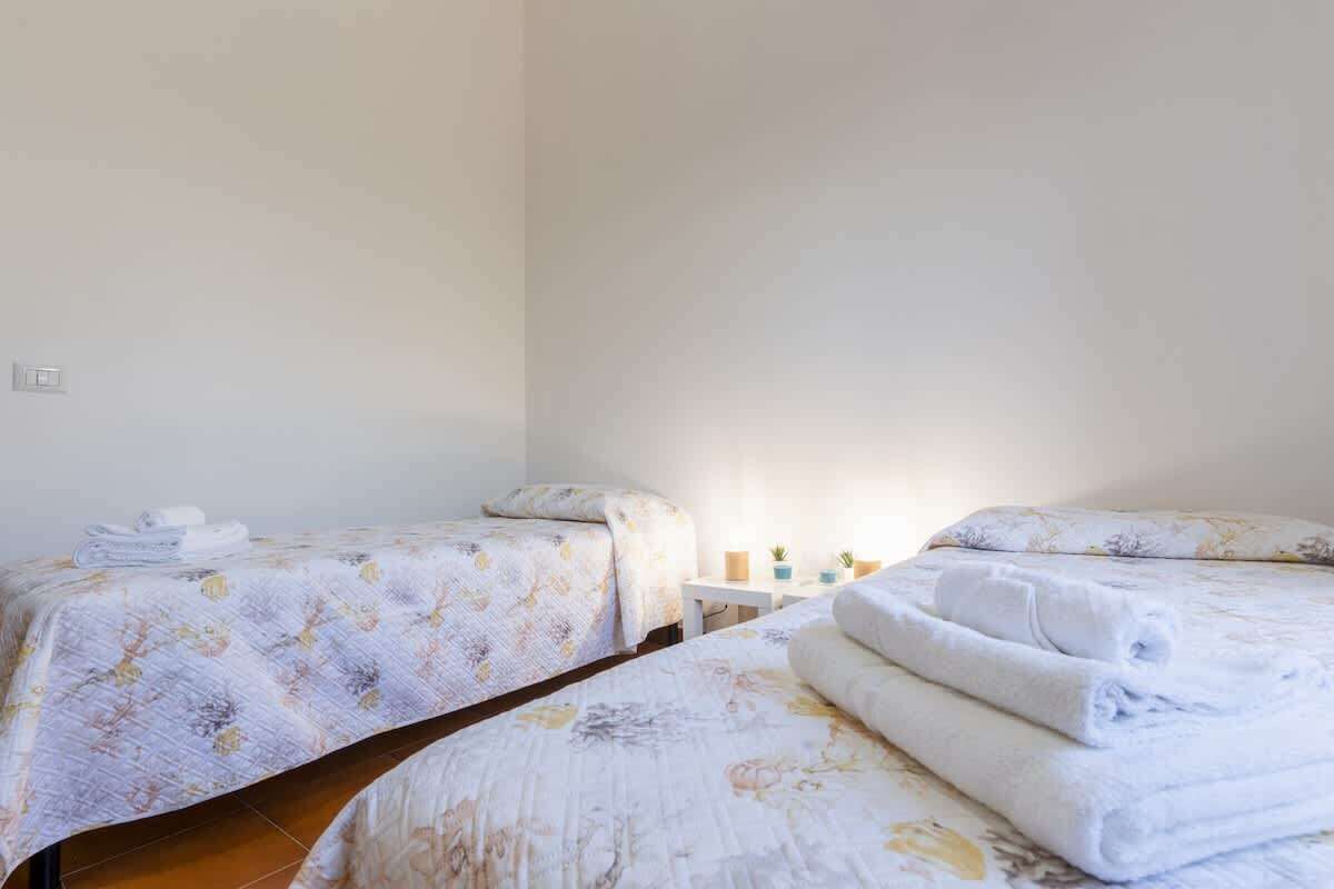A room with two beds, each with a floral bedspread and a neatly folded white towel. There is a small table with a lamp and a few decorative items.