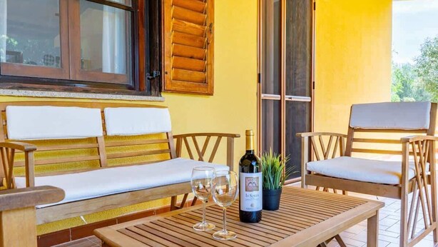 Un banco de madera con cojines blancos, una mesa de madera con dos copas de vino y una botella, y una planta en maceta.