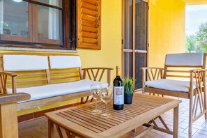 Un banco de madera con cojines blancos, una mesa de madera con dos copas de vino y una botella, y una planta en maceta.