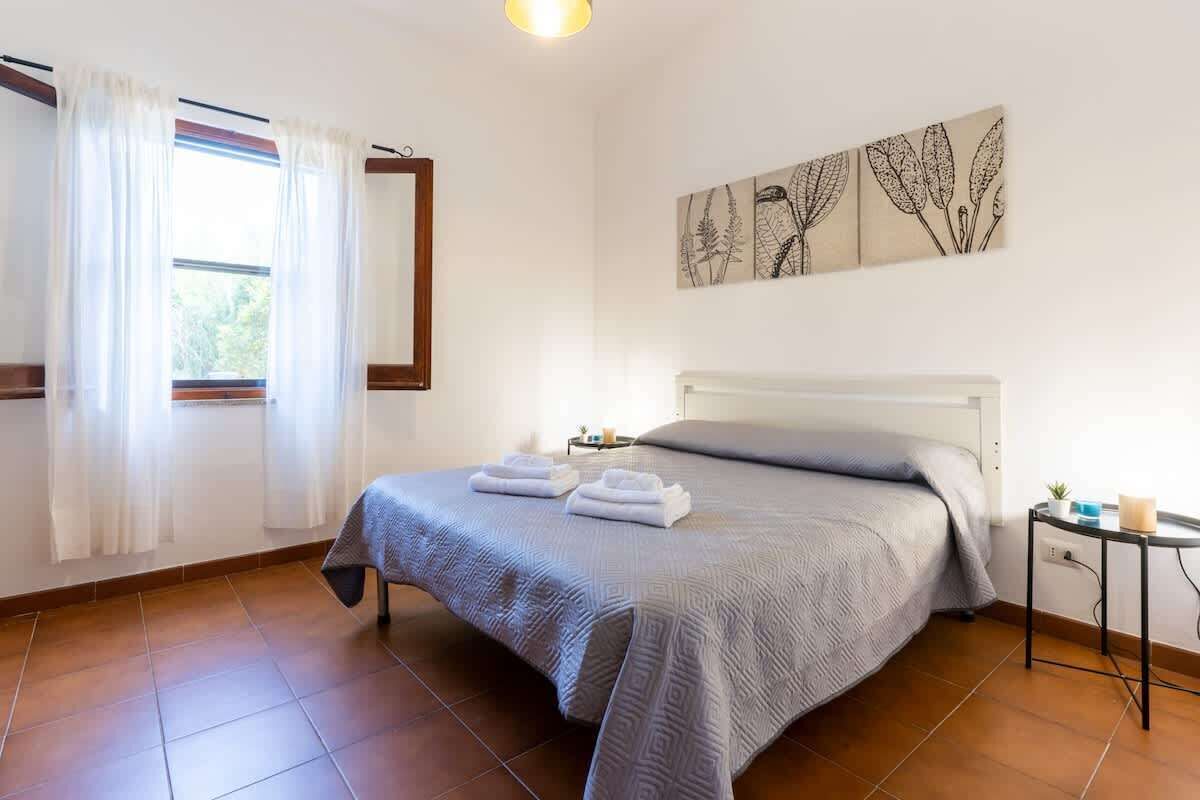 Um quarto com cama, mesa de cabeceira, janela com cortinas e uma obra de arte na parede.