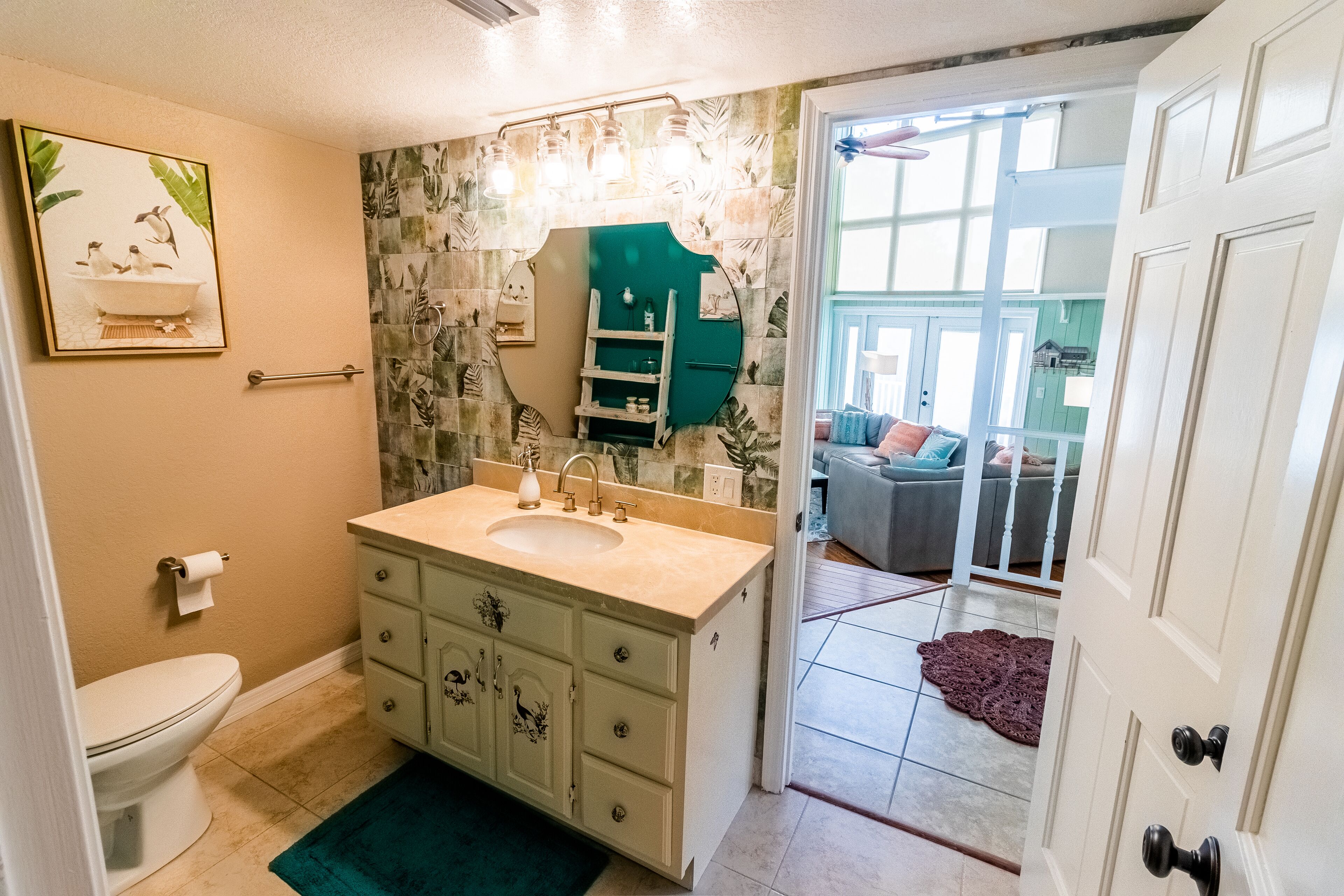 A bathroom with a double sink, a toilet, and a mirror above the sink. There is a door leading to a living area with a couch and a window.