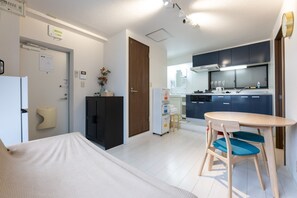 Habitación de hotel pequeña y funcional con cama, kitchenette, comedor con mesa y sillas, y puerta que da a un baño.