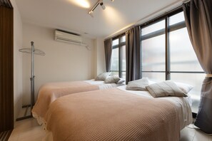 Habitación de hotel con dos camas, un calentador portátil, un equipo de aire acondicionado y ventanas grandes con cortinas.
