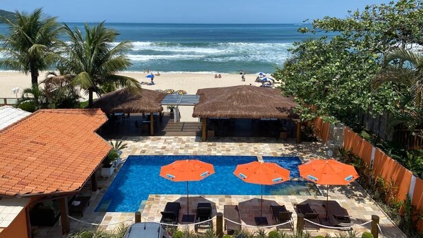 A pool area with lounge chairs and umbrellas, overlooking a beach with people and palm trees. - Pousada Maresias Beira Mar (São Sebastião)