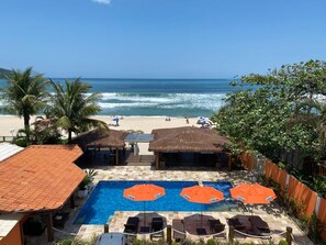 Ein Poolbereich mit Liegestühlen und Sonnenschirmen mit Blick auf einen Strand, an dem sich Menschen und Palmen befinden.