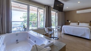 A hotel room with a large window, a bathtub, a bed, a TV, and a table with chairs. - Pousada Maresias Beira Mar (São Sebastião)