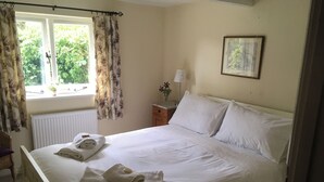 A neatly made bed with towels, a window with floral curtains, a radiator, a bedside table with a lamp, and a framed picture on the wall.