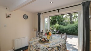 A dining area with a table set for two, a clock on the wall, and a view of the garden through the door.