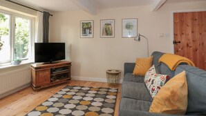 A living room with a TV, wooden entertainment unit, grey sofa with yellow cushions, patterned rug, and a window with curtains.