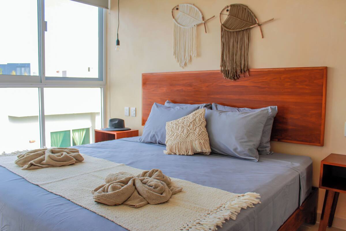 A neatly made bed with a blue and white bedspread, two pillows, and a decorative macrame wall hanging.
