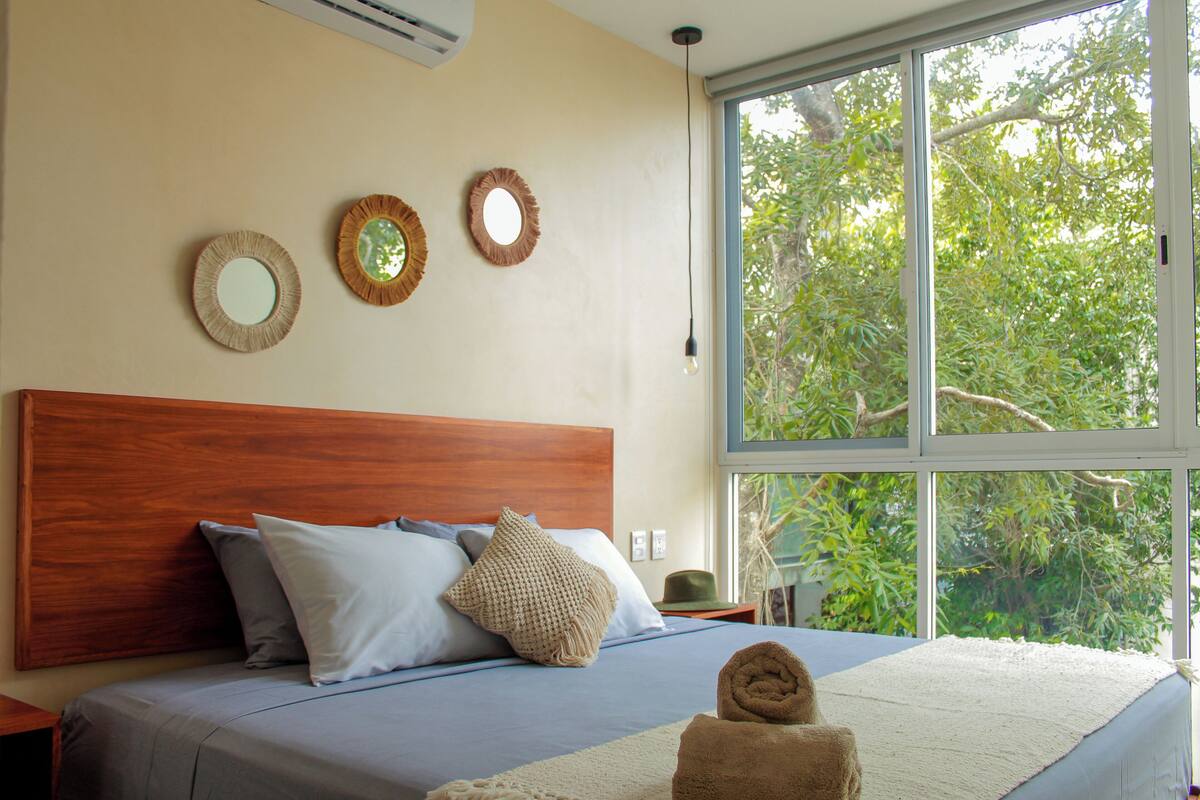 A bedroom with a wooden headboard, a large window with a view of trees, and three wall-mounted mirrors.