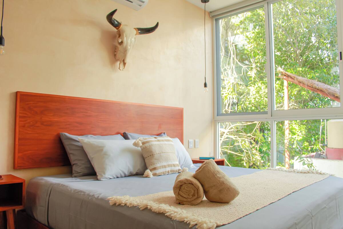 A bedroom with a wooden headboard, a large window with a view of greenery, and a bed with multiple pillows and a rolled towel.